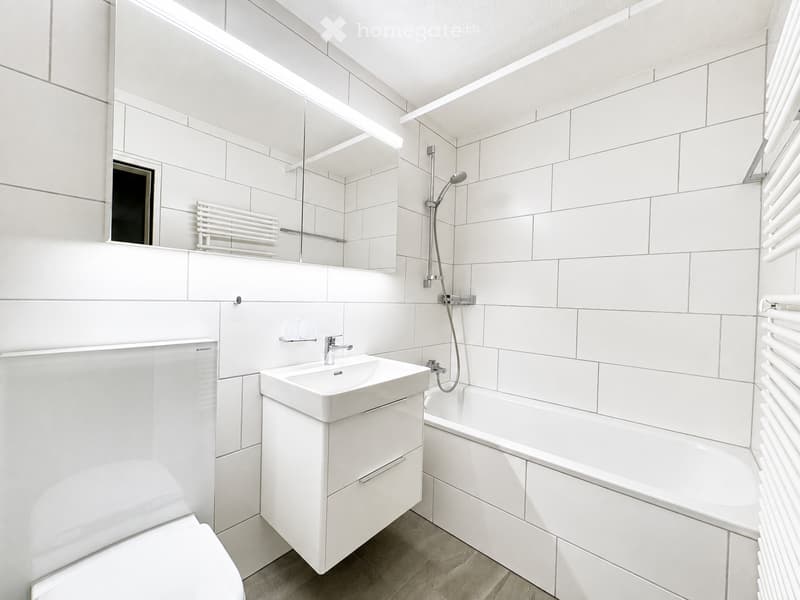 Modern bathroom with white tiles, a sink with a mirror, a bathtub, a toilet, and a radiator. The space features a sleek, contemporary design with built-in storage and a showerhead above the bathtub.