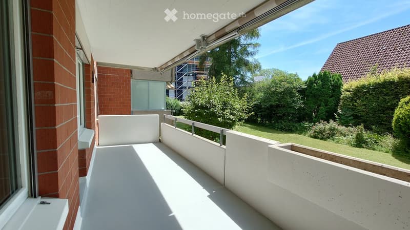 Balcony with white railings, a retractable awning, and a view of a green garden with trees and a neighboring house.