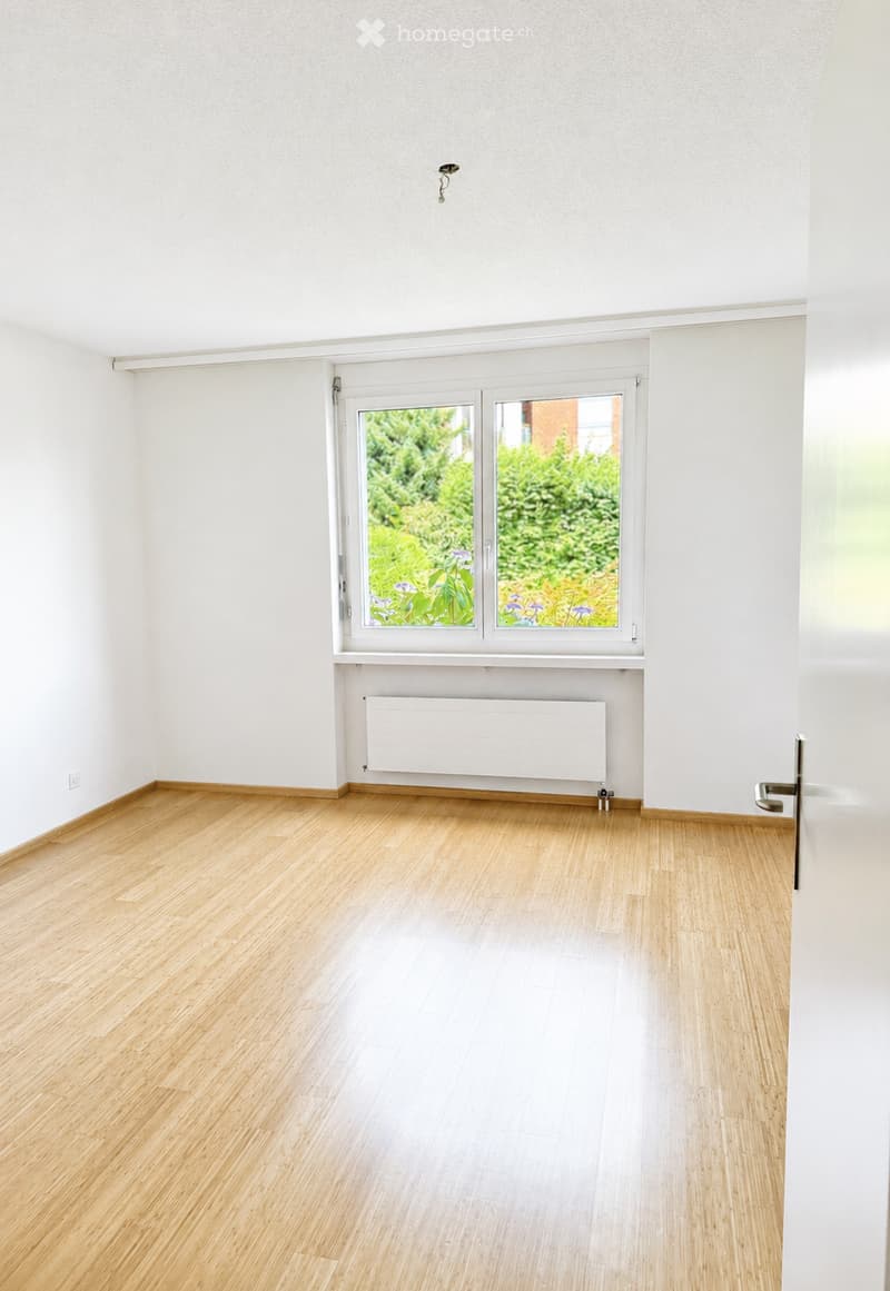 Empty room with light wooden flooring, white walls, a large window with a view of greenery, and a radiator below the window.