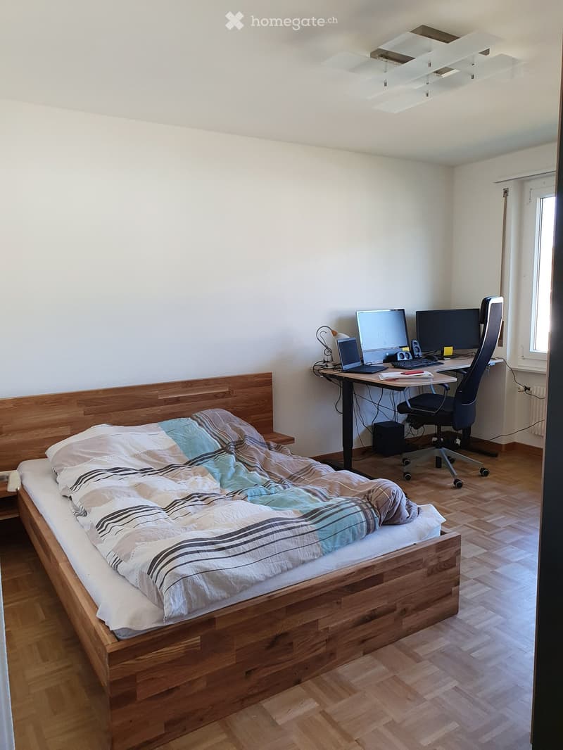 A modern bedroom with a wooden bed, a desk with a laptop and monitors, and a comfortable chair. The room features wooden flooring and large windows allowing natural light.