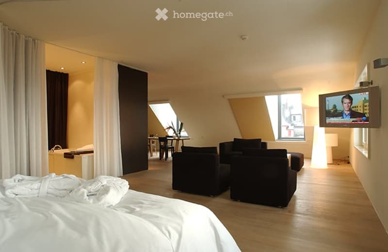 Modern furnished room with a large bed, sofa, chairs, a small table, and a TV screen displaying a program. The room features large windows with a view and warm lighting.
