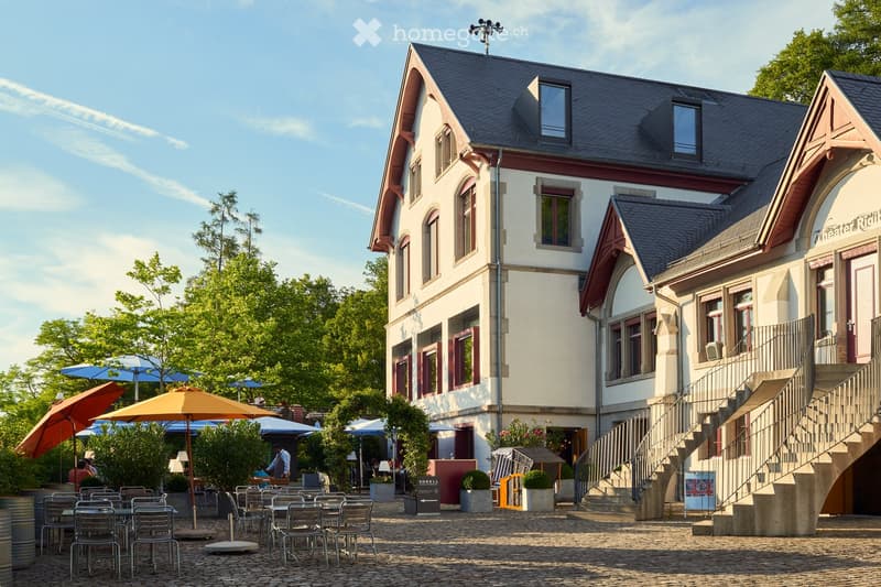 Courtyard area with outdoor seating, umbrellas, and tables, adjacent to a two-story building with white walls, red trim, and multiple windows.