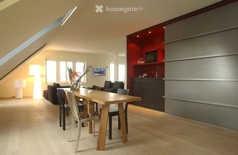 Modern open-plan living and dining area with a wooden dining table, chairs, a sofa, a floor lamp, and a small kitchen area with a counter and sink.