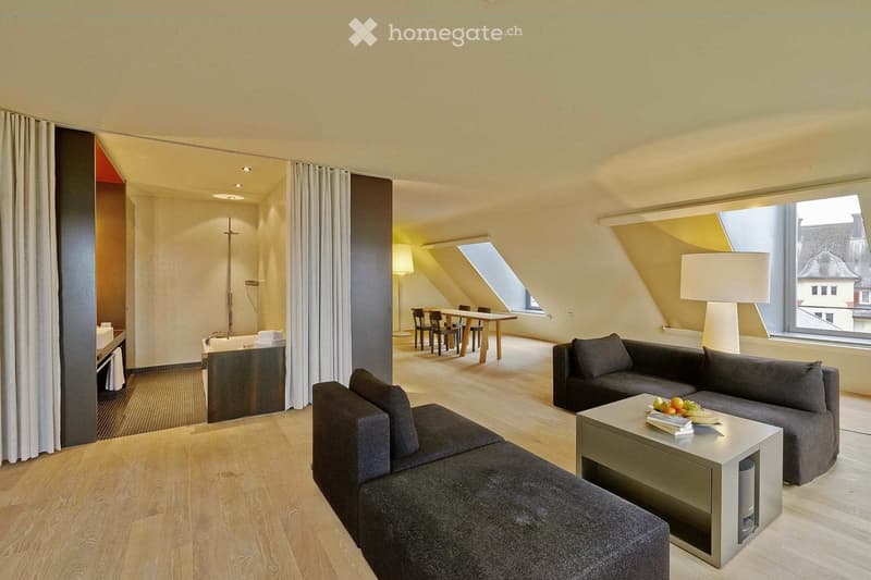 Modern, well-lit living space with a grey sectional sofa, coffee table, dining area with a table and chairs, and a separate bathroom area with a bathtub and sink.
