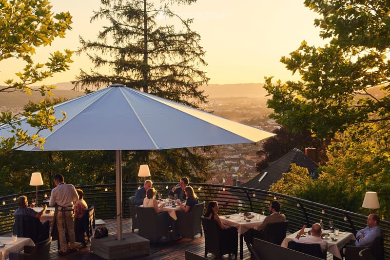 An outdoor terrace with tables and chairs, overlooking a cityscape with trees and a sunset view.