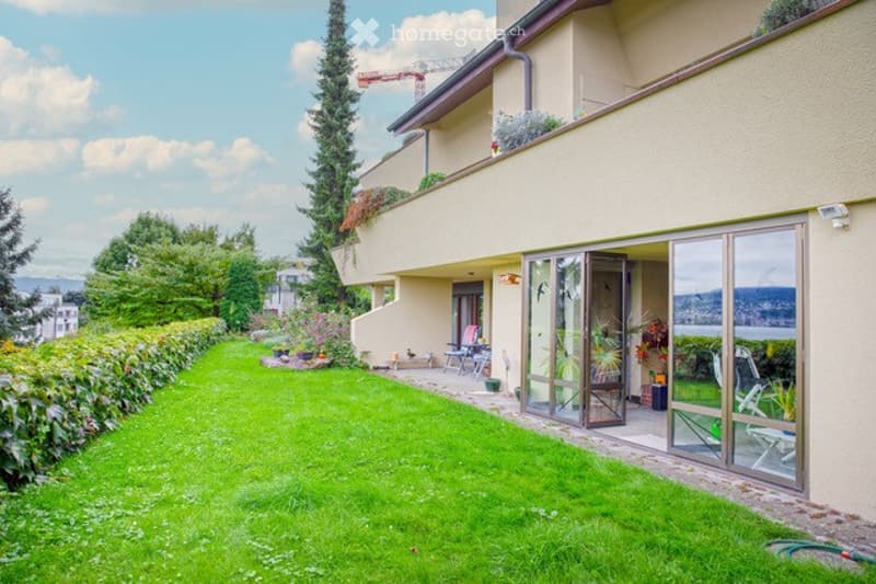 Well-maintained garden with a neatly trimmed lawn, surrounded by trees and shrubs. A modern house with a balcony and large windows overlooks the garden, offering a scenic view of the surrounding landscape.
