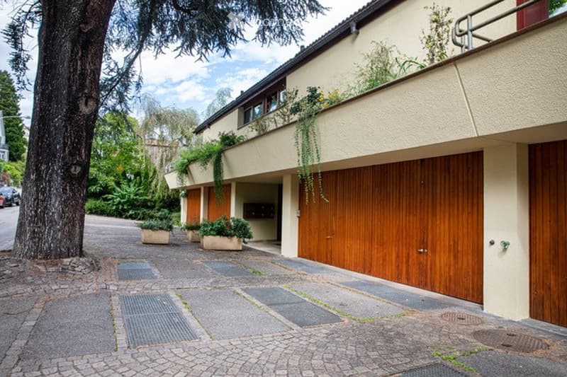 Modern two-story building with a large wooden garage door, balcony with greenery, and a paved driveway.