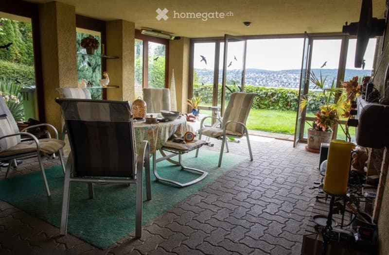 A spacious, well-lit room with large windows offering a scenic view of the landscape. The room features a mix of modern and traditional furniture, including several chairs, a central table, a small stool, and decorative plants and vases.