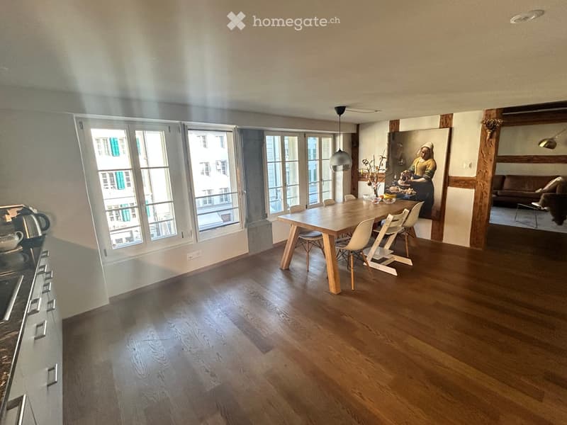 Open-plan living and dining area with wooden flooring, large windows, a dining table with chairs, a kitchen counter, and a sofa in the background.