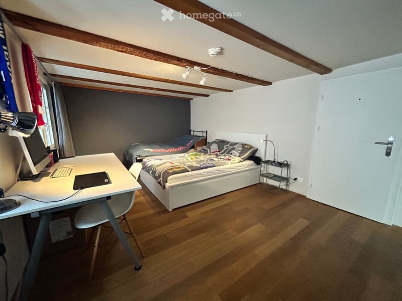 Cozy bedroom with wooden floors, a white bed with colorful bedding, a desk with a laptop and keyboard, and a small nightstand with a lamp.