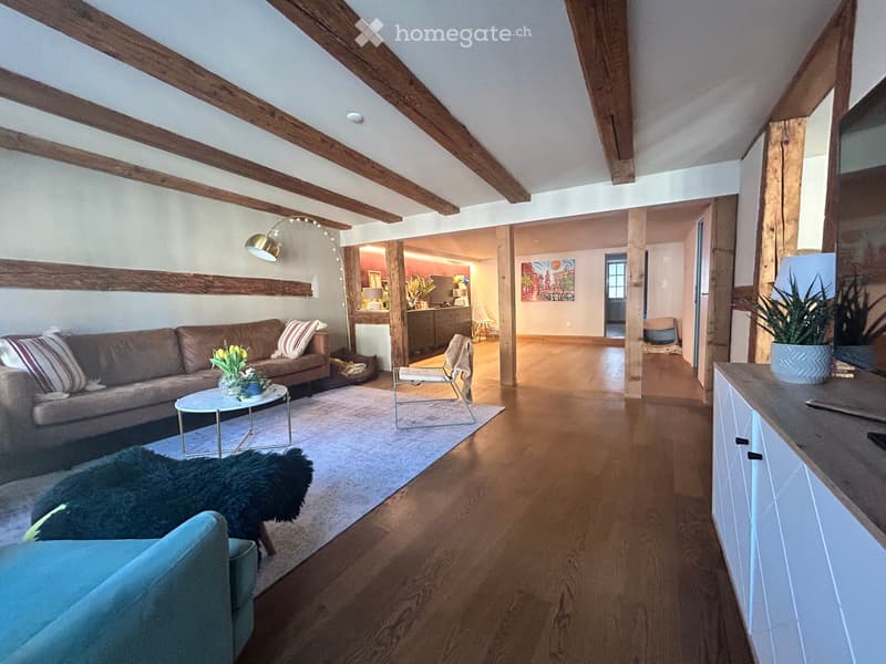 Living room with exposed wooden beams, a brown sofa, a white coffee table, a blue armchair, a fur rug, and a modern pendant light. The room opens to a kitchen area with cabinets and a dining table.
