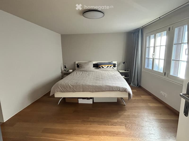 Modern bedroom with wooden flooring, large window with grey curtains, bed with grey bedding, nightstands, and lamps.