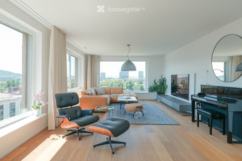 Modern living room with large windows offering a city view, featuring a sectional sofa, coffee table, wicker stool, leather chair, piano, and decorative elements.