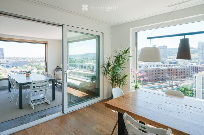 Modern living and dining area with large windows offering a city view, featuring a sofa, coffee table, dining table, chairs, and potted plants.