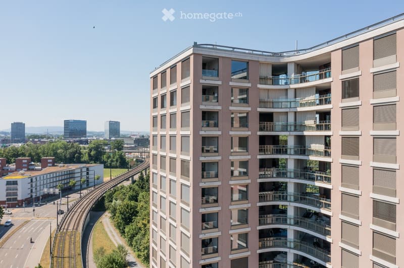 Modern multi-story residential building with balconies, large windows, and contemporary design, set against a cityscape with visible roads, trees, and distant buildings.