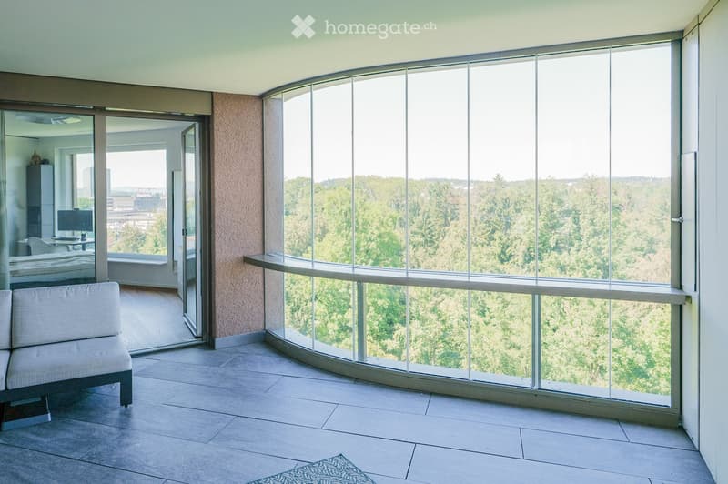 Modern furnished room with large windows offering a scenic view of greenery, featuring a sofa, mirror, table, chair, computer, and bed.
