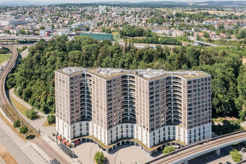 A modern, multi-story residential building with a curved facade, surrounded by greenery and urban infrastructure.