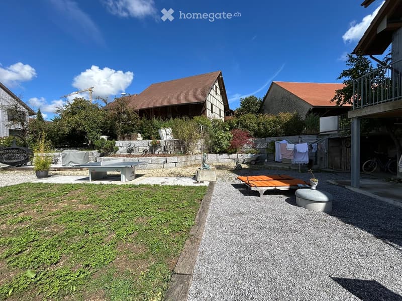 paved seating area with a table, decorative plant, grassy lawn, clothesline with clothes, and stone wall