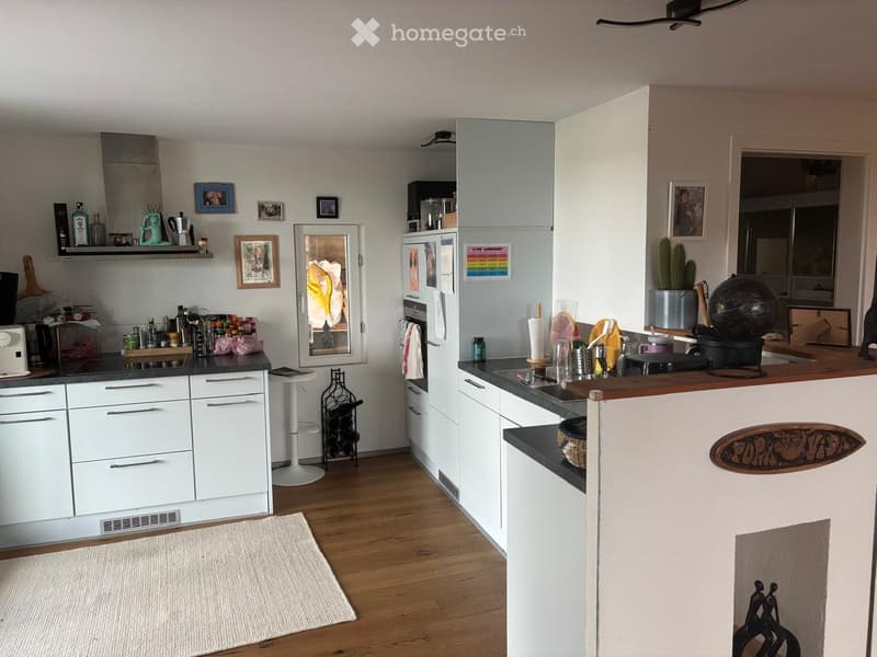 Open kitchen, modern white cabinets, wooden floor, kitchen island with seating, built-in refrigerator and freezer, ample counter space, various kitchen items on countertops, open doorway leading to another room, white tiled walls, window with curtains