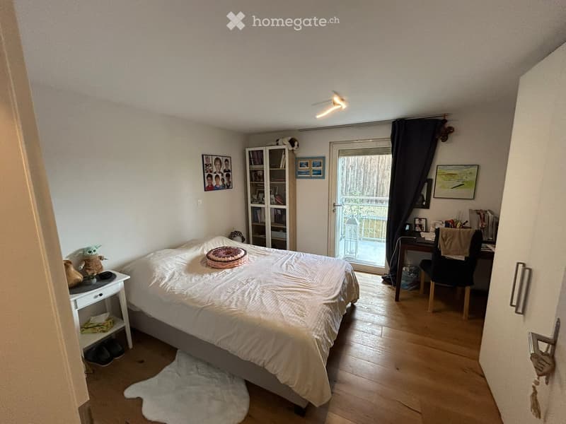 Wooden floor, white walls, bed with white bedding, bedside table with figurines, glass door leading to a balcony, a bookshelf with books and figurines, framed photos on the wall, and a desk by the window. A lamp is mounted on the ceiling. The room has a c