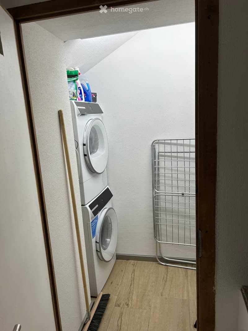 small utility room with stackable washer and dryer, wooden floor, cleaning supplies on wall shelf, collapsible clothes rack