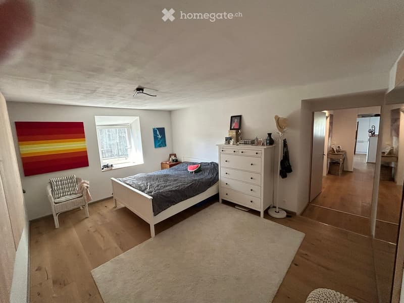 White walls, hardwood floors, bed with grey bedding, a white chair with cushion, a white cabinet with drawers, a rug, colorful painting on wall, open door