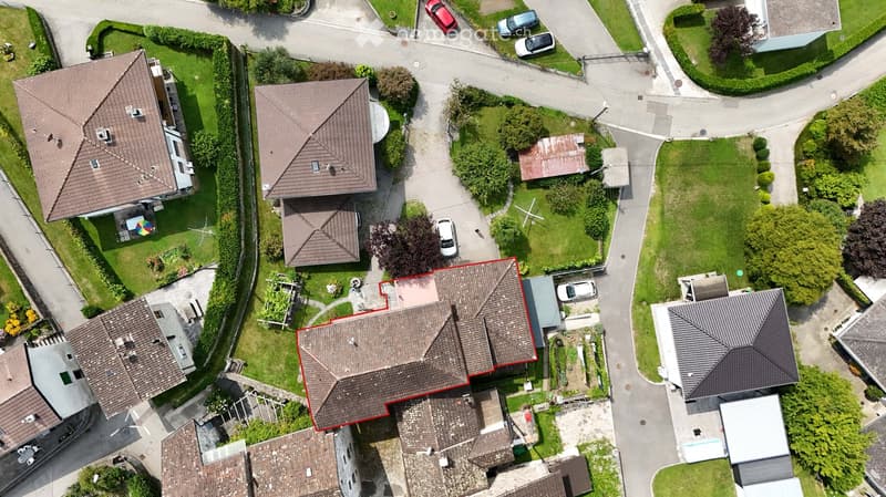 two-story house with brown roof, highlighted in red, surrounded by a well-maintained lawn with various shrubs and trees, and there are cars parked along the road