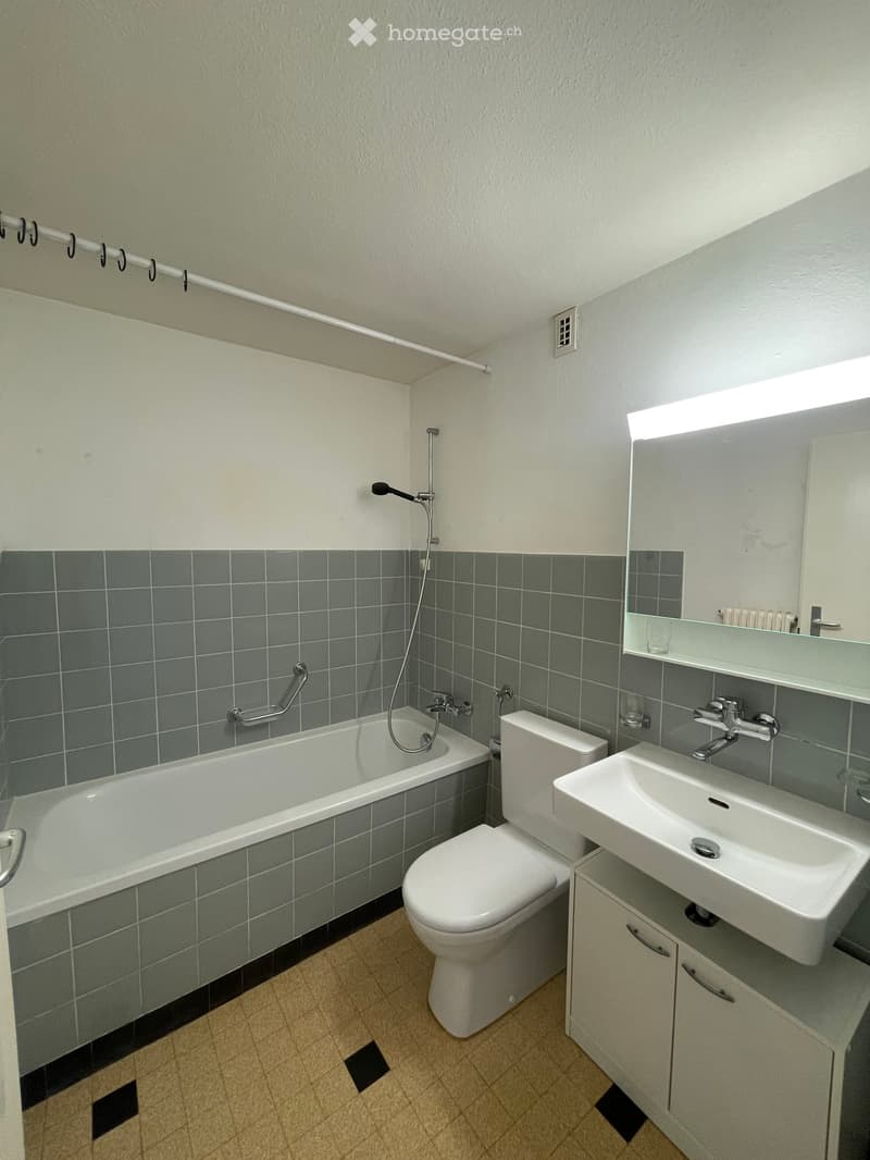 Modern bathroom with a bathtub, toilet, sink, and mirror. Features grey tiled walls and a light-colored floor.