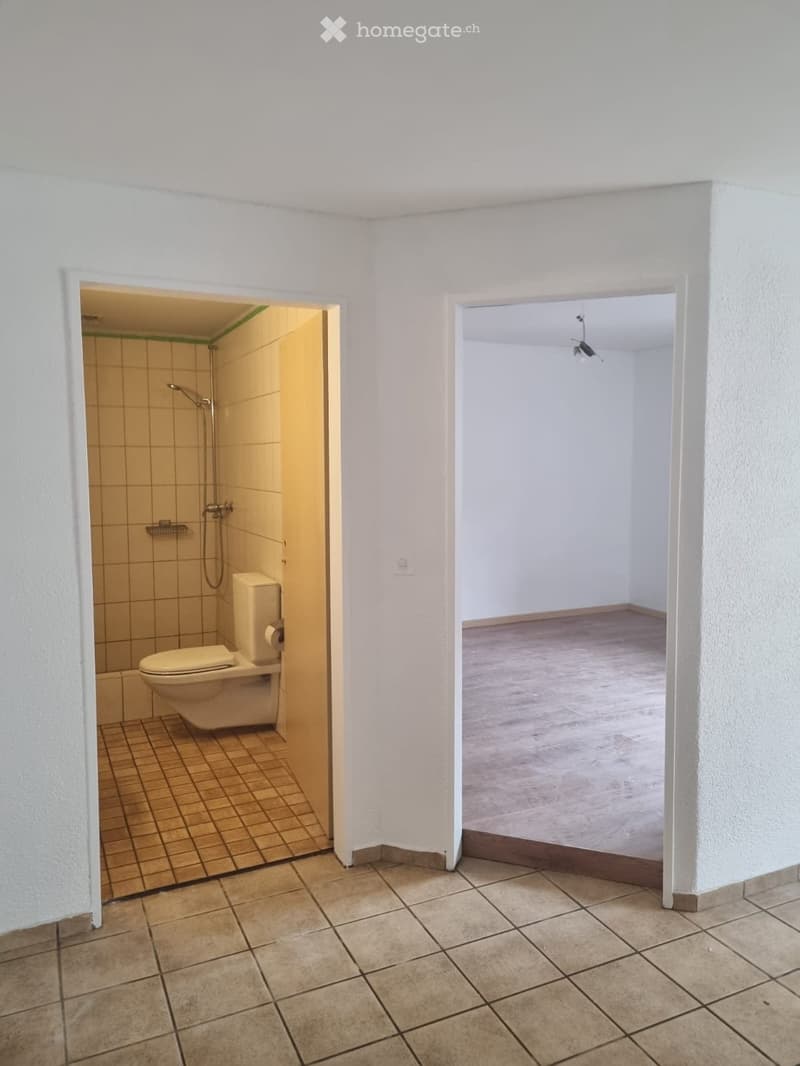 Empty hallway with tiled floor and white walls, leading to a small bathroom with a toilet and shower stall.