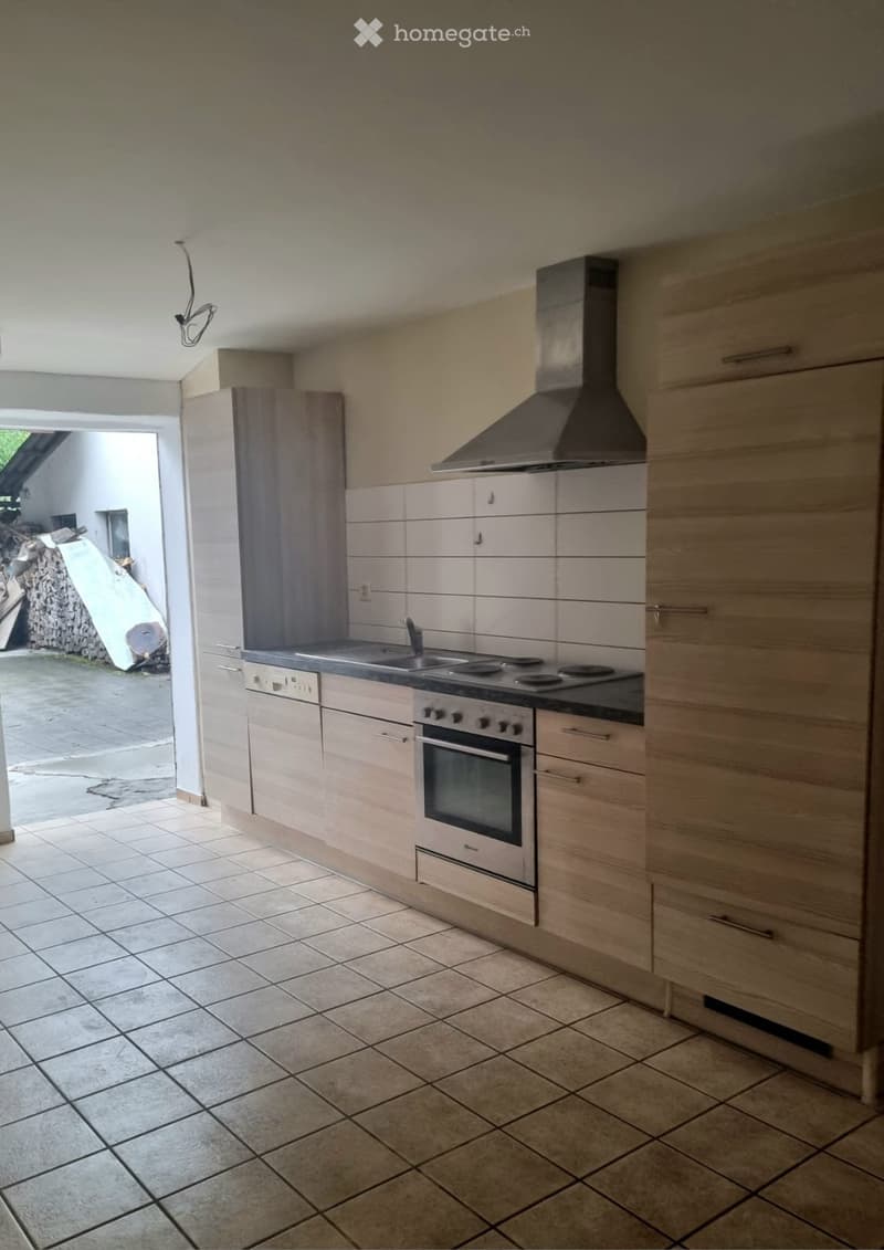 Modern kitchen with light wood cabinets, stainless steel oven, range hood, and tiled floor.