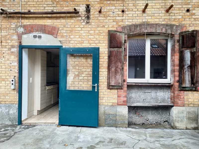 Facade of a brick building with a green door leading to a kitchen area, visible window with wooden shutters, and a small concrete ledge.