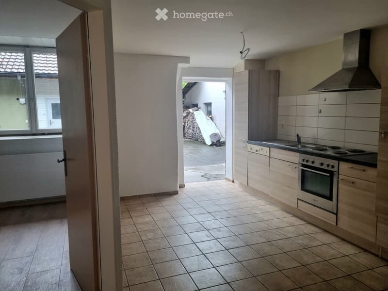 Empty kitchen with tiled floor, light-colored cabinets, oven, sink, and faucet. An open doorway leads to an outdoor area.