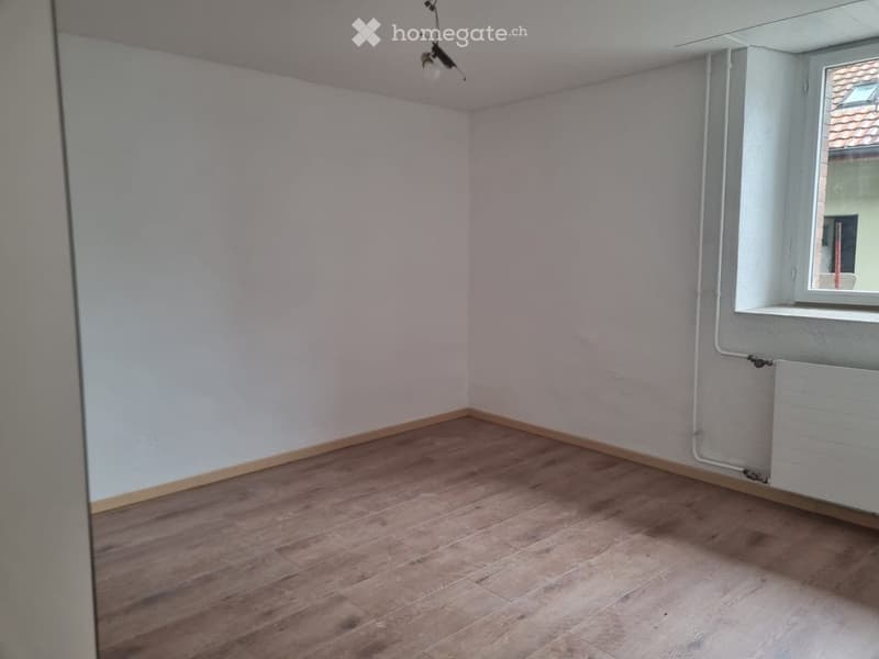 Empty room with light-colored walls, wooden flooring, and a single window with a view of a neighboring building.