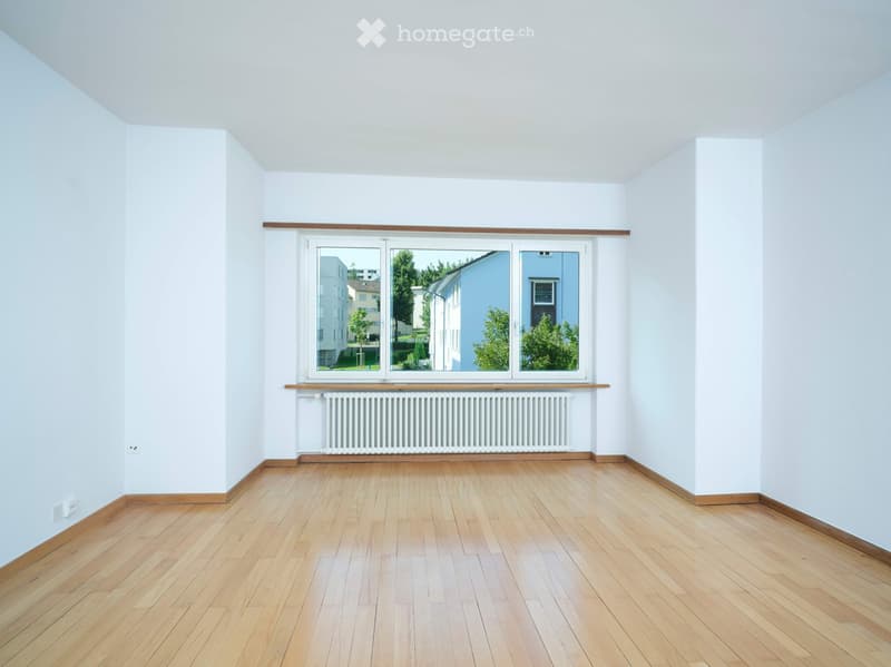 Empty room with light wooden flooring, white walls, and large windows offering a view of the outside. There is a radiator below the windows.