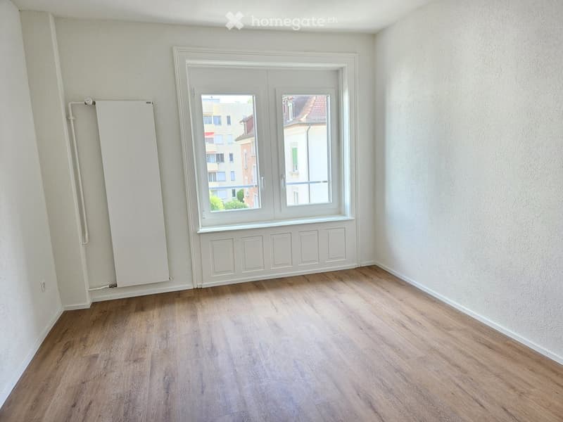 Empty room with wooden flooring, white walls, and a large window with white frame and radiator on the left side.