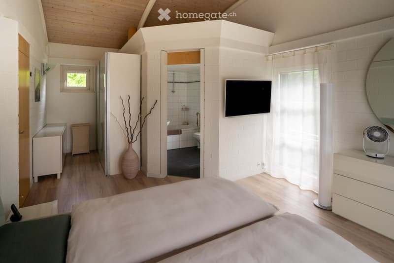 Modern bedroom with a large bed, a flat-screen TV mounted on the wall, a vase with dried branches, a fan on a nightstand, and an open bathroom with a bathtub and toilet.