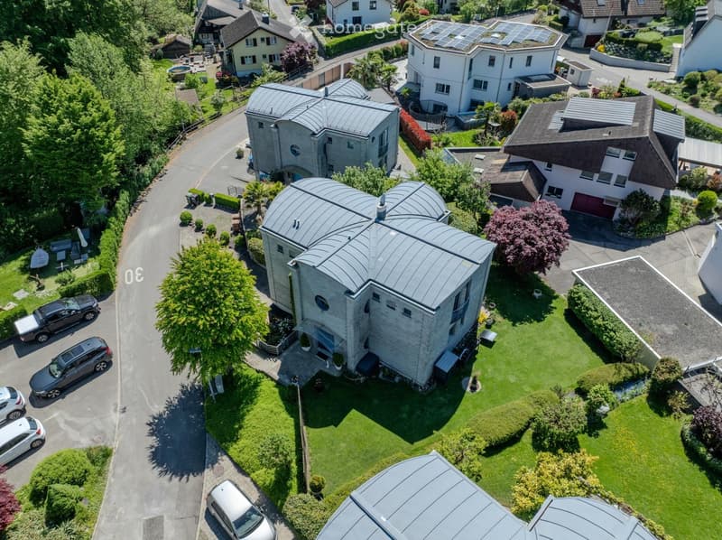 Aerial view of a modern house with a unique curved roof, surrounded by a well-maintained lawn, trees, and neighboring houses. The house features large windows and a driveway with parked cars.