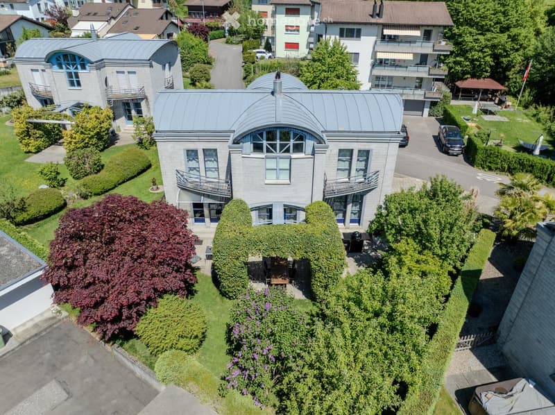 Aerial view of a modern two-story house with grey stone facade, large windows, and a curved metal roof. The house features balconies, a well-maintained garden with trees and shrubs, and is situated in a residential neighborhood with other houses and green