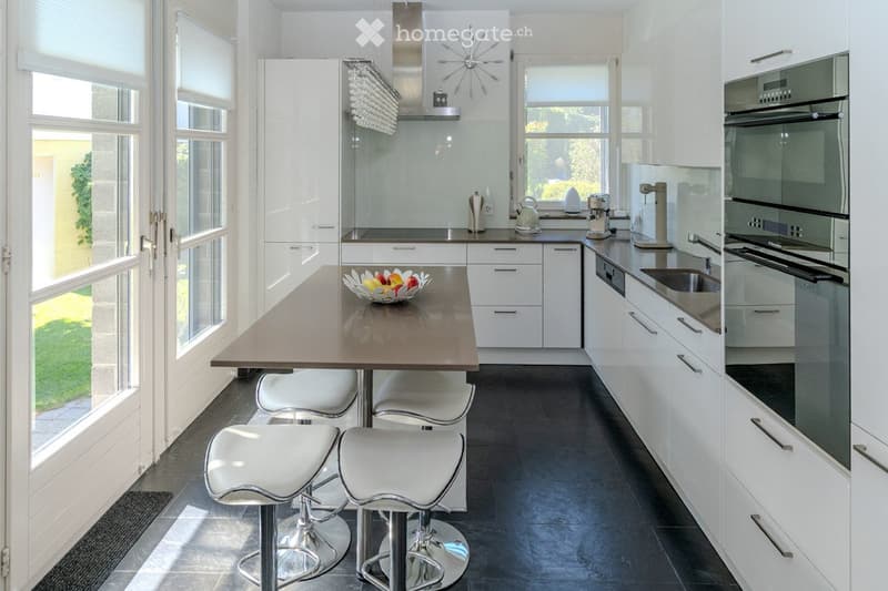 Modern kitchen with white cabinets, stainless steel appliances, island with stools, and a bowl of fruit on the counter.