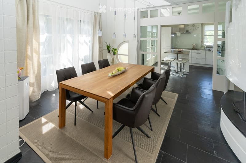 Modern dining area with a wooden table, black chairs, and a small kitchen area with white cabinets and stools.