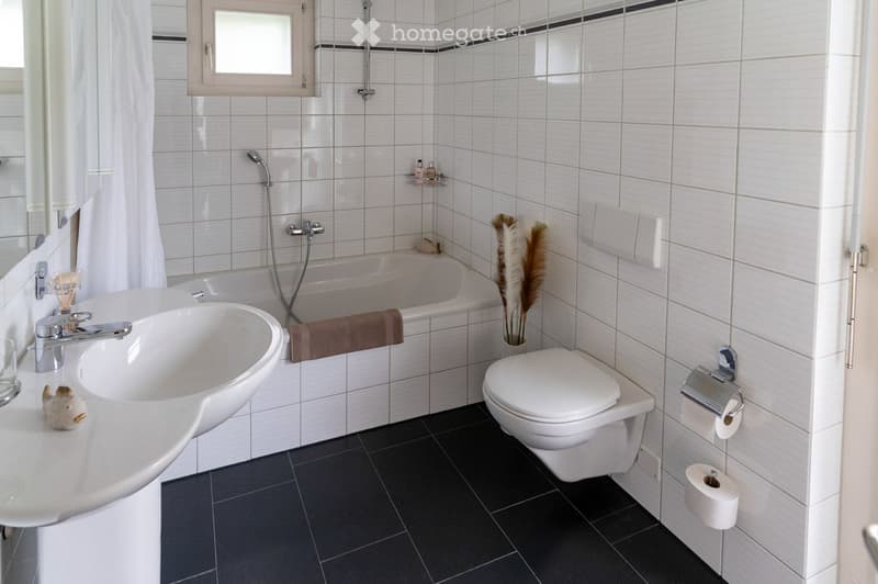 Modern bathroom with white tiles, a bathtub, a sink, a toilet, and a towel on the side. The room features a shower head, a mirror, and a small decorative plant in a pot.