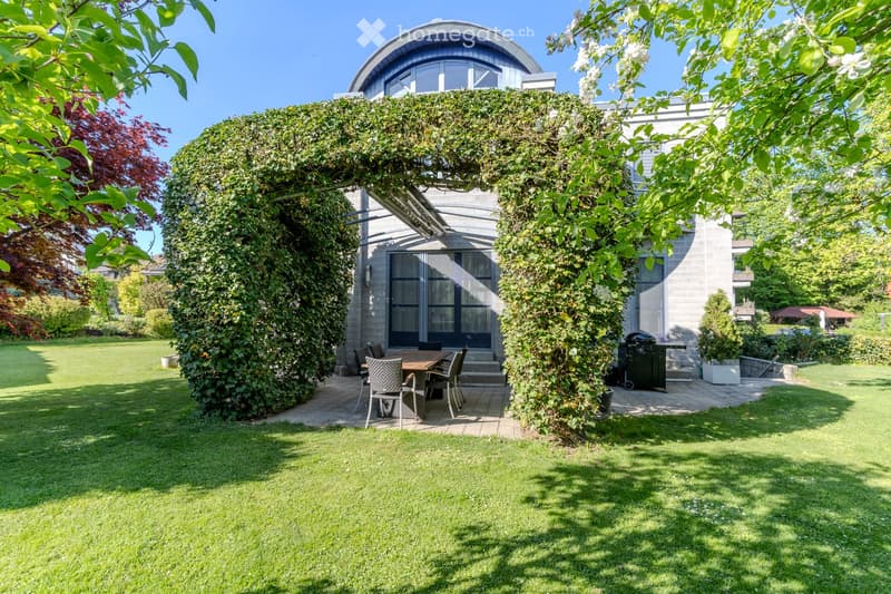 A well-maintained garden with a modern house in the background, featuring a dining set, barbecue, and lush greenery.