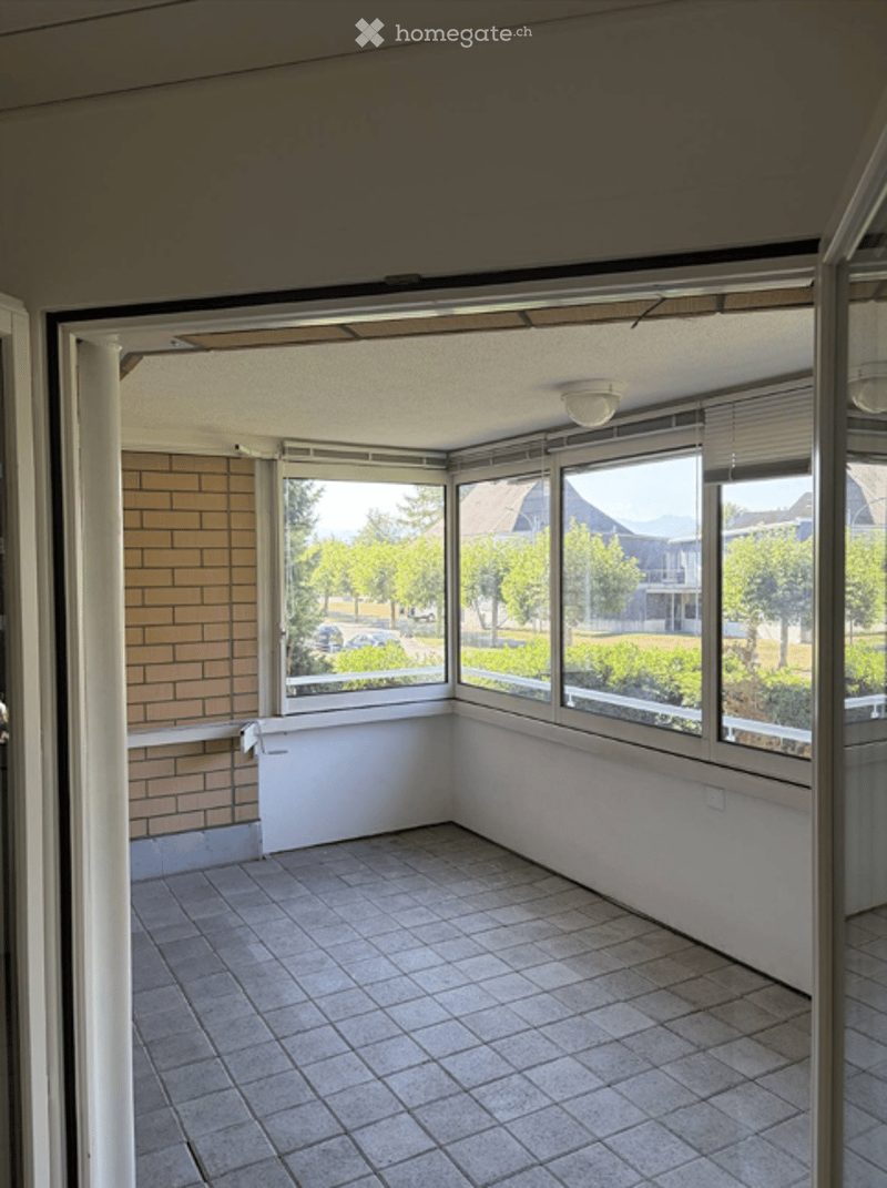 Empty sunroom with tiled floor, large windows, and brick wall. No furniture or equipment present.