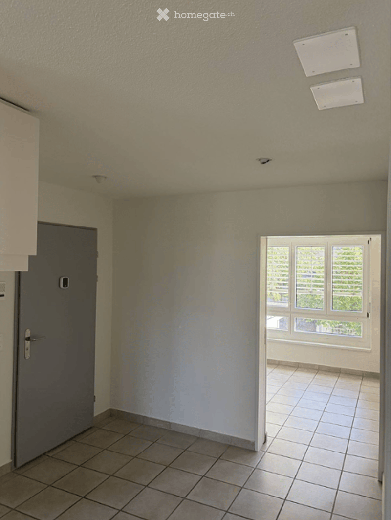 Empty room with tiled floor, white walls, grey door, and an open doorway leading to a balcony area with white tiled flooring and windows.