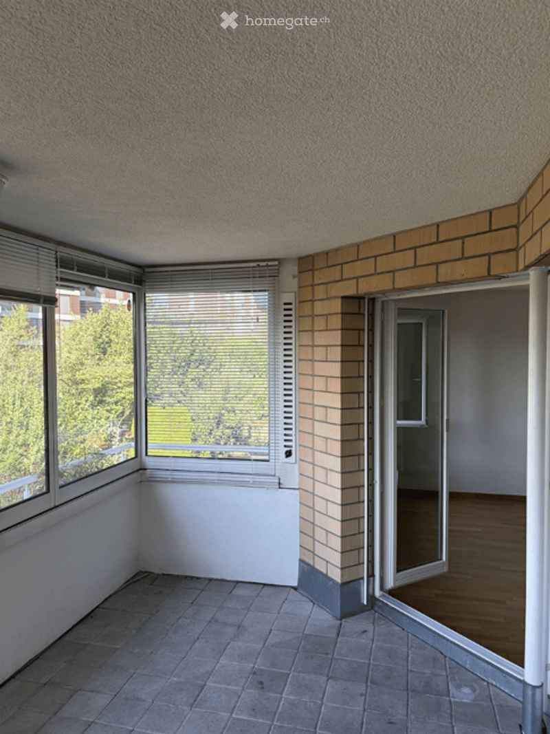 Empty room with tiled floor, large windows with blinds, and an open doorway leading to another room with wooden flooring.