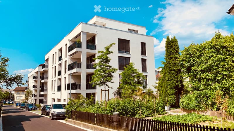 Modern multi-story residential building with white facade, balconies, and surrounding greenery.
