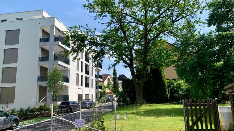 Modern multi-story apartment building with balconies, surrounded by greenery and parked cars.