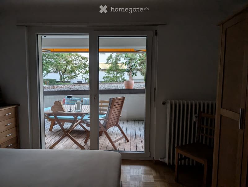 Balcony with wooden floor, table, chairs, and a radiator visible inside the room.