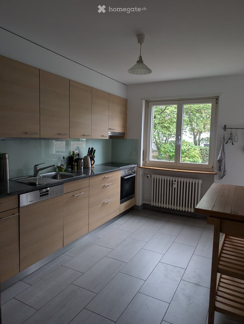 Modern kitchen with light wooden cabinets, granite countertop, sink, oven, dishwasher, and a small wooden table with a chair.