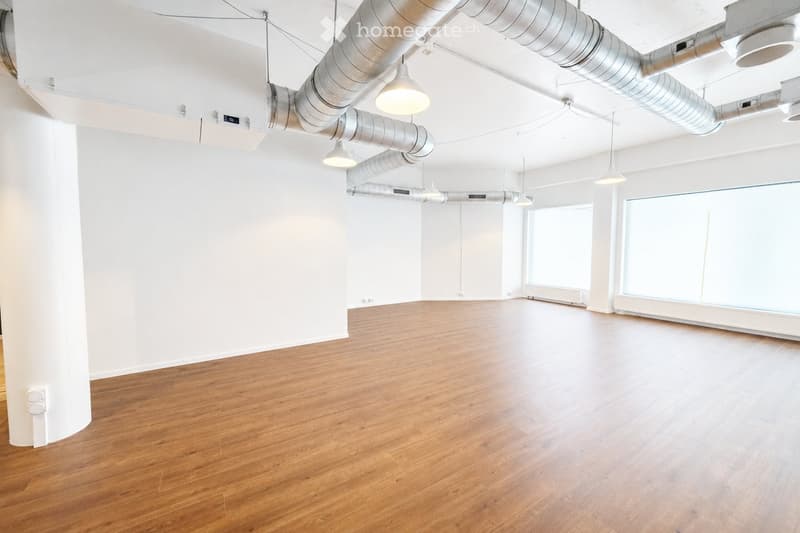 Empty room with wooden flooring, white walls, large windows, and exposed ductwork with hanging lights.